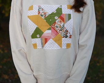 May include: Cream-colored sweatshirt featuring a patchwork design. The square patch showcases a starburst pattern with floral, geometric, and dotted fabric elements in shades of yellow, green, pink, and white. The sweatshirt has long sleeves and a crew neck.