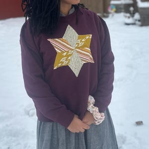 Puede incluir: Una sudadera morada oscura con una gran aplicación de estrella multicolor en la parte delantera. La estrella presenta secciones de telas doradas, rosas y blancas. La persona lleva una falda gris y una goma para el pelo estampada en la muñeca.