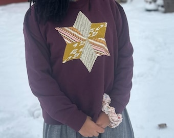 Könnte beinhalten: Ein dunkelvioletter Pullover mit einem großen, mehrfarbigen Stern-Applikation auf der Vorderseite. Der Stern hat Abschnitte aus goldenen, rosa und weißen Stoffen. Die Person trägt einen grauen Rock und hat eine gemusterte Haargummi am Handgelenk.