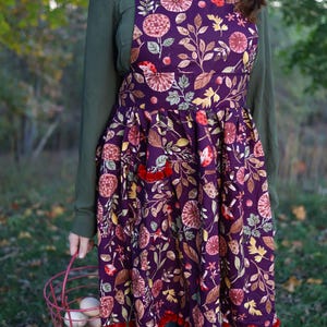 Puede incluir: Un delantal floral morado con ribete de borlas rojas. El delantal presenta un estampado botánico con flores y hojas en tonos rojos, amarillos y marrones. Se sostiene una cesta roja llena de huevos.