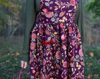 Könnte beinhalten: Ein lila Blumenschürze mit rotem Bommelbesatz. Die Schürze hat einen botanischen Druck mit Blumen und Blättern in Rot-, Gelb- und Brauntönen. Ein roter Drahtkorb mit Eiern wird gehalten.