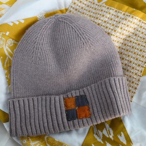 May include: A light brown knit beanie with a ribbed brim and a square patch. The patch is made of two squares, one dark blue and one orange, stitched together. The hat is on a yellow and white patterned quilt.