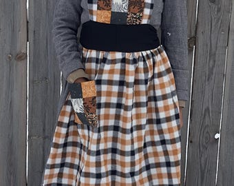 Könnte beinhalten: Eine handgefertigte Schürze mit einem Patchwork-Latz und einer Tasche, mit einem schwarz-weiß-braunen Karomuster-Rock. Die Schürze hat einen schwarzen Bund und wird über einem grauen Kleid getragen. Im Hintergrund ein Holzzaun.