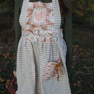 Puede incluir: Delantal a rayas beige y blanco con un diseño de patchwork con un corazón rosa y motivos florales. El delantal tiene un bolsillo con un lazo decorativo y está diseñado para uso en la cocina o el jardín.