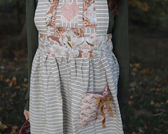 Könnte beinhalten: Beige-weiß gestreifte Schürze mit Patchwork-Design mit einem rosa Herzen und floralen Mustern. Die Schürze hat eine Tasche mit einer dekorativen Schleife und ist für die Küche oder den Garten konzipiert.