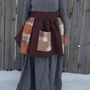 Puede incluir: Un delantal marrón con bolsillos de retazos y paneles decorativos. El delantal se usa sobre un vestido y una camisa grises. El patchwork incluye patrones naranjas, azules y marrones. El fondo es una valla de madera y nieve.