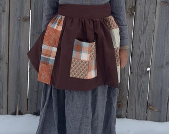 Könnte beinhalten: Eine braune Schürze mit Patchwork-Taschen und dekorativen Einsätzen. Die Schürze wird über einem grauen Kleid und Hemd getragen. Der Patchwork enthält orange, blaue und braune Muster. Der Hintergrund ist ein Holzzaun und Schnee.