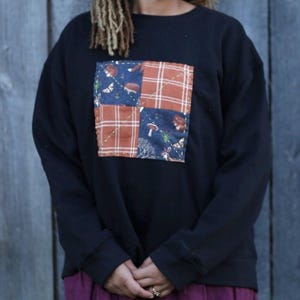 Puede incluir: Sudadera negra con dise&ntilde;o de patchwork. El patchwork presenta cuadrados de tela azul con criaturas del bosque y cuadros naranjas. La sudadera tiene mangas largas y cuello redondo. La persona lleva una falda morada.