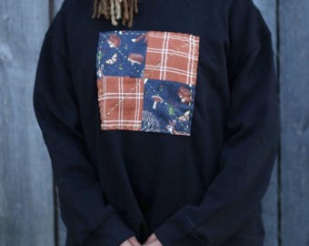 May include: Black sweatshirt with a patchwork design. The patchwork features squares of blue fabric with woodland creatures and orange plaid. The sweatshirt has long sleeves and a crew neck. The person is wearing a purple skirt.