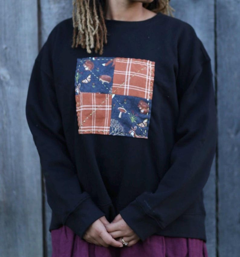 Puede incluir: Sudadera negra con dise&ntilde;o de patchwork. El patchwork presenta cuadrados de tela azul con criaturas del bosque y cuadros naranjas. La sudadera tiene mangas largas y cuello redondo. La persona lleva una falda morada.