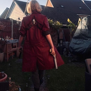May include: A long, deep red coat featuring a structured collar, shoulder panels, and a metallic skull emblem centered on the upper back. The garment is worn with dark trousers and includes two long, silver-toned handheld pistols held at the sides.