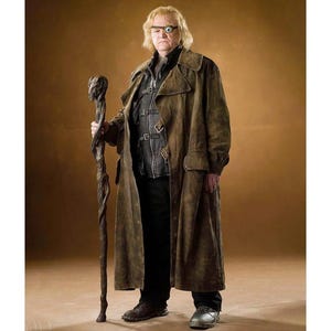 Puede incluir: Un hombre con un abrigo largo marrón y pantalones negros, sosteniendo un bastón de madera. Tiene un ojo protésico y lleva un chaleco de cuero. El fondo es de color marrón liso.