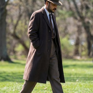 May include: A man in a brown overcoat, trousers, and a fedora hat walks outdoors. The coat is buttoned, and the man wears a white shirt and brown tie. Black shoes complete the outfit. The background is a park with trees and grass.