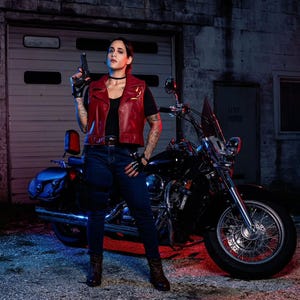 May include: A person wearing a dark red leather vest over a black t-shirt, dark blue jeans, and fingerless gloves, holding a black handgun. Beside them is a black cruiser motorcycle with chrome accents, parked in front of a closed garage door.