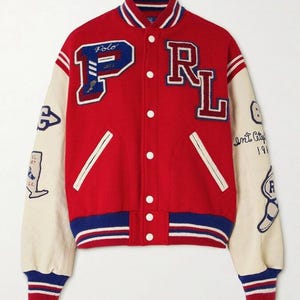 Op de afbeelding: Rode varsity jas met crèmekleurige mouwen. De jas heeft de letters "PRL" in collegestijl, met geborduurde patches. De mouwen hebben blauw-rood gestreepte manchetten. De jas heeft een knoopsluiting.