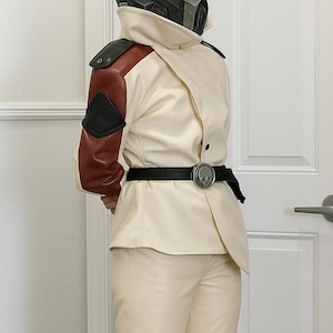 Könnte beinhalten: Cremefarbenes futuristisches Kostüm mit einem schwarzen und roten Akzent auf der linken Schulter. Das Outfit umfasst einen passenden Helm, eine Jacke, eine Hose und einen schwarzen Gürtel mit silberner Schnalle. Die Jacke hat ein einzigartiges asymmetrisches Design.