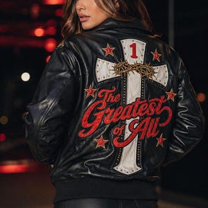 Pode incluir: Casaco bomber de couro preto com um design de cruz branca nas costas, com detalhes de estrelas vermelhas e o texto "The Greatest Of All". O número "1" está acima da cruz, e uma coroa de espinhos dourada está no centro.