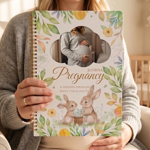 Puede incluir: Un diario de embarazo con encuadernación en espiral que presenta una ilustración en acuarela de dos conejos, hojas verdes y flores amarillas. La portada muestra el texto Pregnancy Journal: A Modern Pregnancy Guide for Moms to Be y una foto.