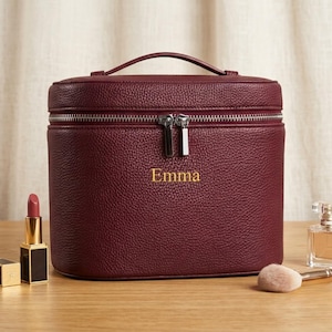 May include: A burgundy pebbled leather vanity case with a top handle and a silver double zipper closure. The name Emma is embroidered in gold thread on the front. A lipstick and a perfume bottle are positioned on the surface next to the case.