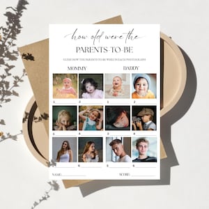 以下が含まれることがあります： 「how old were the PARENTS-TO-BE」と「GUESS HOW THE PARENTS-TO-BE WERE IN EACH PHOTOGRAPH」というテキストが入った白いカード。カードには「MOMMY」と「DADDY」の写真スペースがあり、各写真の下には番号が記載されています。カードはベージュの皿の上にあります。