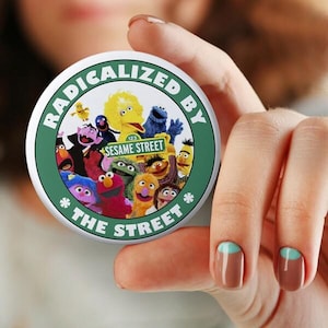 May include: A round button with a green border and the text "RADICALIZED BY THE STREET". The center features Sesame Street characters including Big Bird, Elmo, and Cookie Monster. The button is held in a hand.
