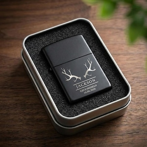 Può includere: Un accendino nero con un design a corna d'argento, il nome "Jackson" e il testo "Groomsman 16.06.2026" sono visualizzati. L'accendino è in una scatola di latta argentata con imbottitura in schiuma nera.