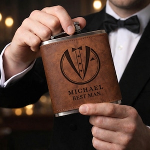 Puede incluir: Una petaca de cuero marrón con una tapa de acero inoxidable. La petaca presenta un gráfico de esmoquin y el nombre "MICHAEL BEST MAN" grabado en la parte delantera. La petaca la sostiene una persona que lleva un traje negro y una pajarita.