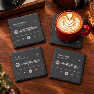 May include: Four square black slate coasters featuring white printed music player interfaces. Three coasters display song titles and artist names: Invisible String by Taylor Swift, My Girl by The Temptations, and Booster Seat by Spacey Jane.