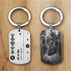 Può includere: Due portachiavi argentati. Uno presenta un design a tema musicale con un codice Spotify e il testo "All of Me" di John Legend. L'altro portachiavi ha una foto in bianco e nero di una coppia che si bacia.