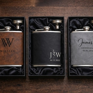 Può includere: Tre fiaschette personalizzate in scatole nere con fodera in raso nero. Le fiaschette sono marroni, nere e grigie, ciascuna con un tappo argentato e un piccolo imbuto. La fiaschetta marrone dice "William", la fiaschetta nera ha le iniziali e la fiaschetta grigia dice "James, Groomsman 06.06.2016".