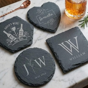 May include: A selection of slate coasters in different shapes, including square, round, and heart. The coasters have engraved designs, such as initials, names, and wine-related graphics. The coasters are dark grey with white engraved text.