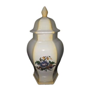 May include: A white ceramic jar with a lid, featuring a floral design in blue, yellow, and brown. The jar has a hexagonal base and a light yellow accent on the sides and lid. The finial on the lid is also light yellow.