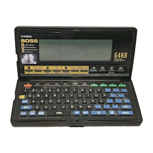 May include: A black Casio BOSS 64KB digital organizer with a flip-top screen and a QWERTY keyboard. The device features a monochrome display, yellow function keys, and blue letter keys. Text on the unit includes CASIO, BOSS, and 64KB SUPER MEMORY.