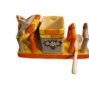 Könnte beinhalten: Ein Vintage-Salz- und Pfefferstreuer-Set aus Keramik. Das Set besteht aus einem rechteckigen Behälter mit floralem Design, einem kleinen Löffel und einem dekorativen Element. Die Farbpalette umfasst Gelb, Orange, Grau und Weiß.