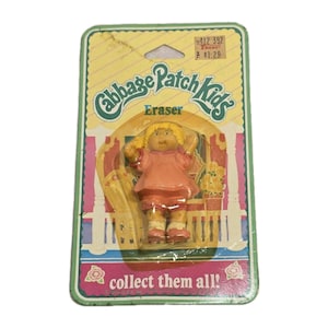 Puede incluir: Una goma de borrar de Cabbage Patch Kids en su embalaje original. La goma es una figura de muñeca con pelo rubio y un vestido rosa. El embalaje es una tarjeta verde y amarilla con las palabras "Cabbage Patch Kids" y "collect them all!"