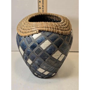 Puede incluir: Un jarrón decorativo de cerámica con borde tejido. El jarrón presenta un patrón geométrico de cuadrados azules, blancos y grises. El borde está hecho de fibras naturales muy tejidas. El jarrón tiene una forma y un diseño únicos.