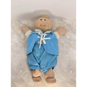 Op de afbeelding: Een vintage Cabbage Patch Kid pop, met een zacht, bruin gezicht, blauwe ogen en een kleine pluk blond haar. De pop draagt een lichtblauwe outfit, inclusief jas en broek, vastgemaakt met een witte strik.