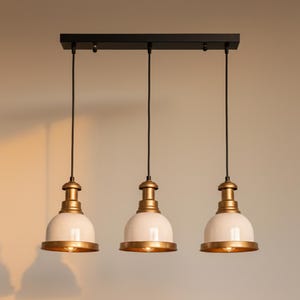 May include: Three-light pendant fitting with a black rectangular base. Each light has a white, bell-shaped shade with a gold-coloured rim and accents. The lights hang from black cords.