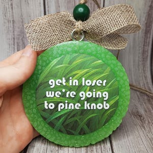 May include: A round, green air freshener with the text "get in loser we're going to pine knob." The air freshener has a grass design in the center and a textured border. It is adorned with a burlap bow and a green bead.