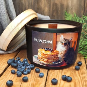 Pode incluir: Uma vela preta com tampa de madeira e um rótulo com um gato e panquecas com mirtilos. O rótulo diz "YOU BETCHA!". Os mirtilos estão espalhados na superfície de madeira.