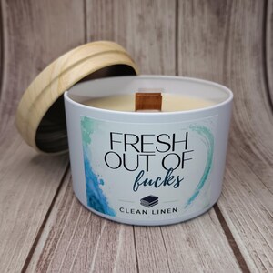 May include: A white candle in a metal tin with a wooden lid. The label features blue and green watercolor accents and the text "FRESH OUT OF fucks" above a small icon of stacked books and the words "CLEAN LINEN" in black lettering.