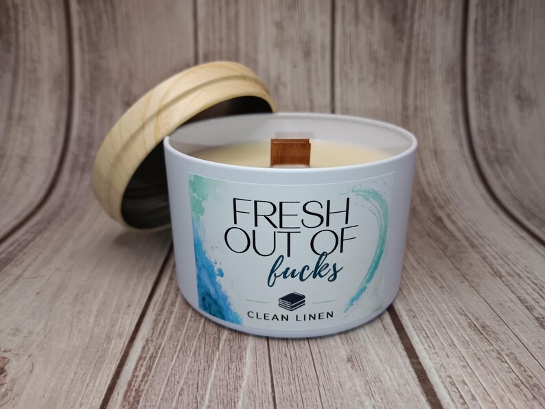 May include: A white candle in a metal tin with a wooden lid. The label features blue and green watercolor accents and the text "FRESH OUT OF fucks" above a small icon of stacked books and the words "CLEAN LINEN" in black lettering.
