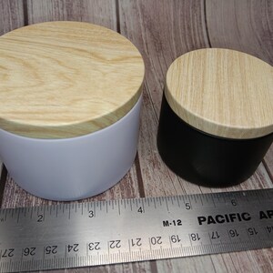 May include: A white cylindrical container and a smaller black cylindrical container, both featuring light-colored wood grain lids. A metal ruler is positioned in front, showing the white container is approximately 3.5 inches in diameter.