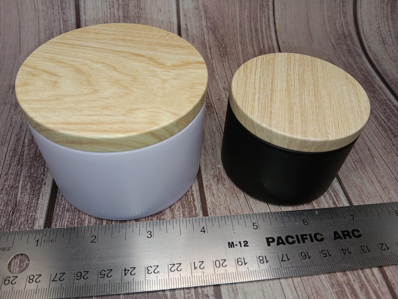 May include: A white cylindrical container and a smaller black cylindrical container, both featuring light-colored wood grain lids. A metal ruler is positioned in front, showing the white container is approximately 3.5 inches in diameter.