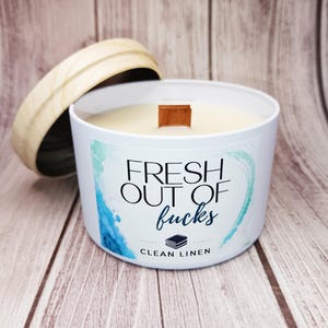 May include: A white candle in a metal tin with a wooden lid. The label features blue watercolor accents and the text "FRESH OUT OF fucks" in black and blue, with "CLEAN LINEN" printed below a small icon of a stack of books and a wooden wick.