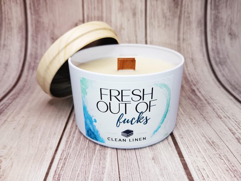 May include: A white candle in a metal tin with a wooden lid. The label features blue watercolor accents and the text "FRESH OUT OF fucks" in black and blue, with "CLEAN LINEN" printed below a small icon of a stack of books and a wooden wick.