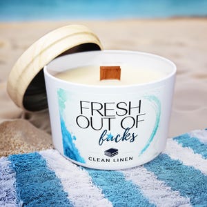 May include: A white candle in a round container with a wooden lid. The label features blue watercolor accents and the text "FRESH OUT OF facks" in black and blue script, with "CLEAN LINEN" and a small stack of books icon printed below.