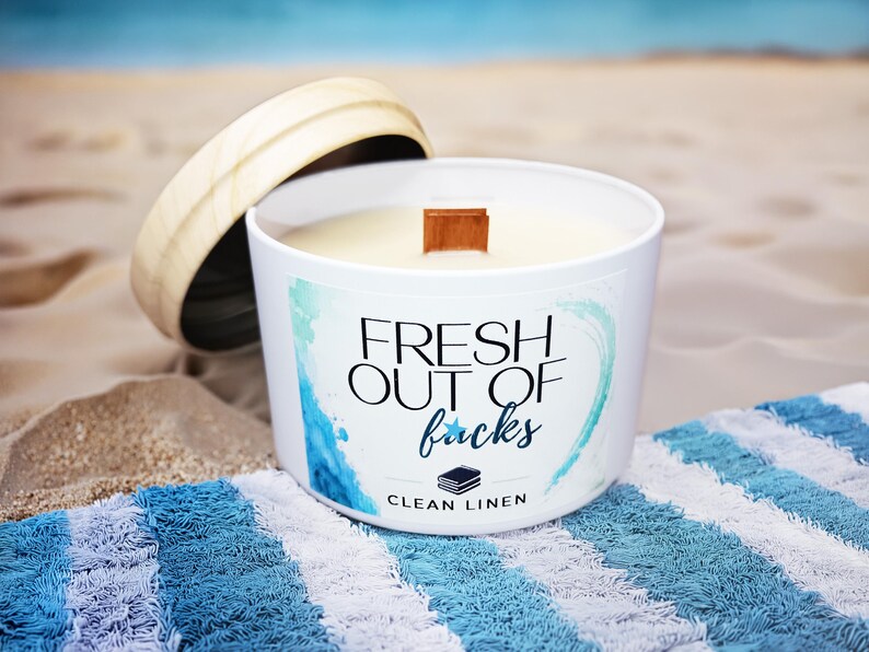 May include: A white candle in a round container with a wooden lid. The label features blue watercolor accents and the text "FRESH OUT OF facks" in black and blue script, with "CLEAN LINEN" and a small stack of books icon printed below.