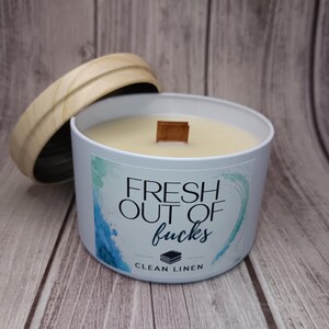 May include: A white metal candle tin with a wooden lid and a wooden wick. The label features blue and green watercolor accents and the text "FRESH OUT OF fucks" above a small icon of folded linens and the words "CLEAN LINEN" in black lettering.