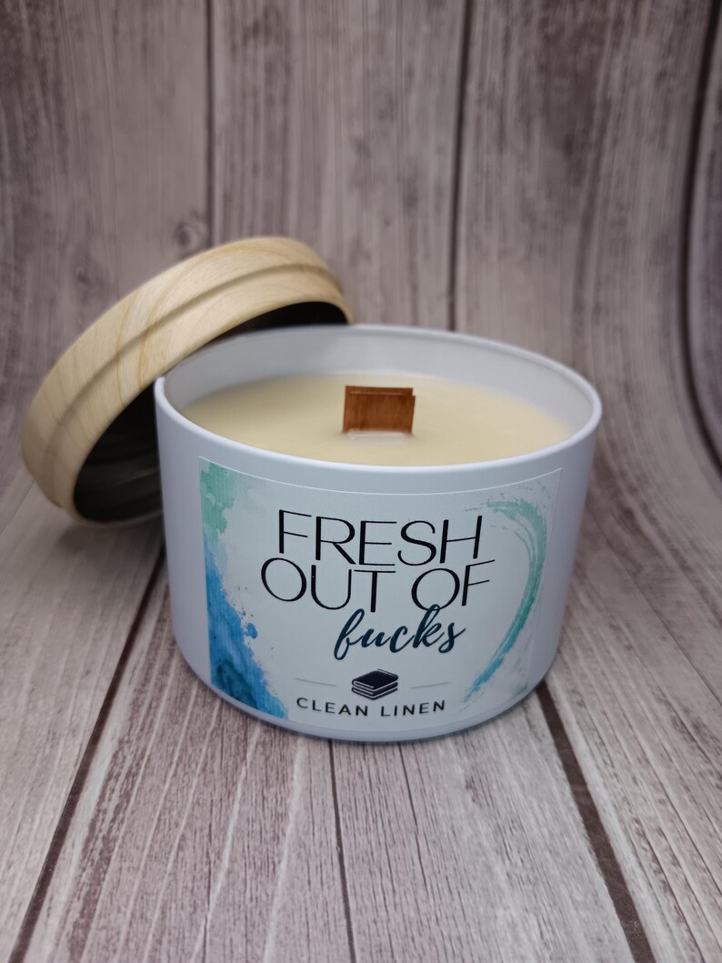 May include: A white metal candle tin with a wooden lid and a wooden wick. The label features blue and green watercolor accents and the text "FRESH OUT OF fucks" above a small icon of folded linens and the words "CLEAN LINEN" in black lettering.